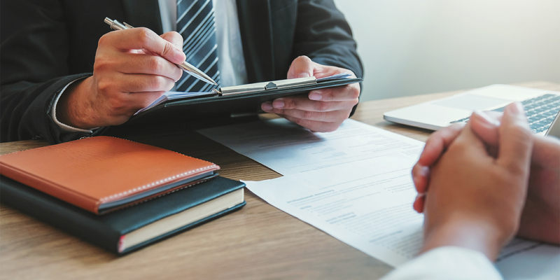 A man in a suit with a clipboard speaking to a female across the table with her hands held together on a table of documents. Court of Appeal affirms extended role of solicitors in assisting catastrophically injured clients