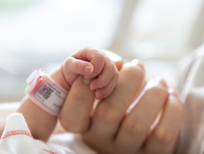 A newborn baby's arm and hand with identification band on its wrist, holding an adult hand. Continuing the conversation around birth trauma and childbirth-related PTSD