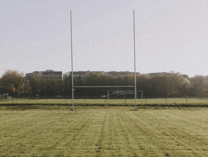 An image of a rugby pitch in a park. Rugby goal posts are directly in front and approximately 30 meters away. There are tress in the background and four flats flat roofs visible beyond.