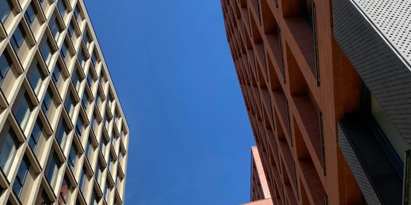 Clear blue sky framed by two modern buildings with geometric window patterns, one in beige tones and the other in reddish-brown tones.